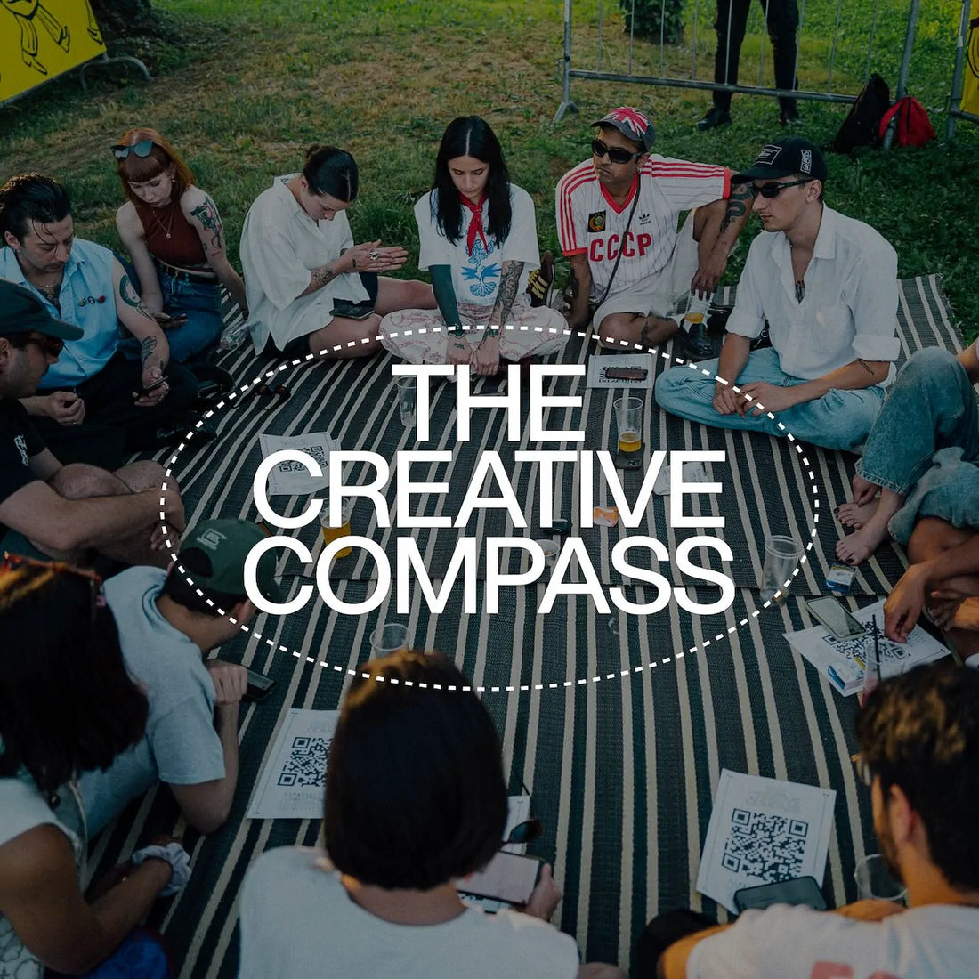 The foreground shows the white "The Creative Compass" logo while on the background, people are sitting around a big towel laying on the grass